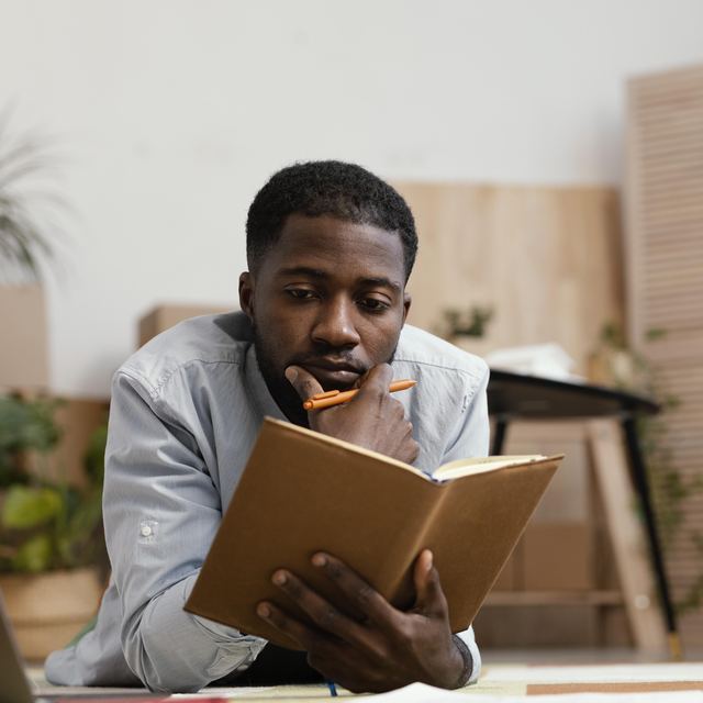 A man lying on a living-room floor, reading a notebook with a pencil in hand.